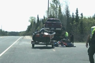 Trailer with damaged snowmobile, tow vehicle, and gear on roadside.