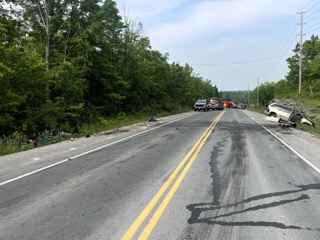 Vehicle wreckage and emergency responders on a two-lane road.