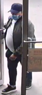 Person wearing a black baseball cap and face mask, black jacket, white shirt, and black pants. Holding a brown paper bag.