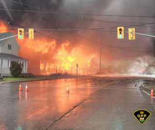 Large house engulfed in flames, orange smoke fills the sky, rain on the street, traffic cones visible