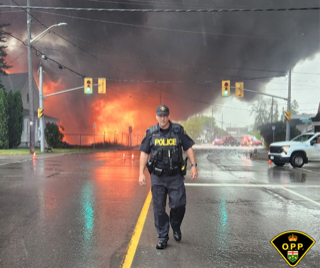 Police officer walking down a wet street, fire and smoke in the background.