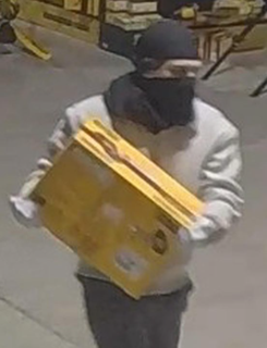 Person wearing a dark hooded sweatshirt and black face covering carries a yellow box.