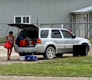White SUV with open tailgate, filled with red containers. Person standing nearby wearing a red top and shorts.