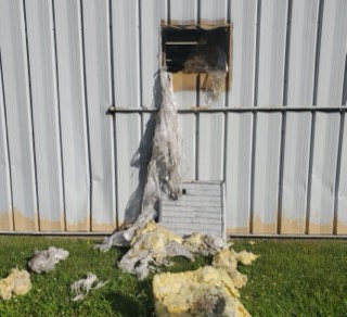 White insulation and plastic sheeting hanging from a hole in a corrugated metal wall.