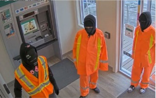 Three people in hooded sweatshirts and reflective orange work vests stand near an ATM.