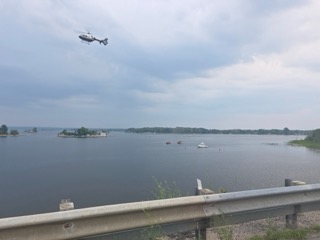 Helicopter flying over a lake, boats visible, shoreline in the distance