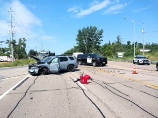 Damaged gray SUV, debris on road, police vehicles, roadside vegetation