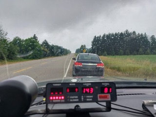 Dashboard display showing 43 in the left display and 81 in the right display. Black car ahead on a two-lane road. Green trees lining the road.