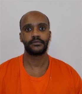 Man with a shaved head and beard, wearing an orange t-shirt, looking directly at the camera.