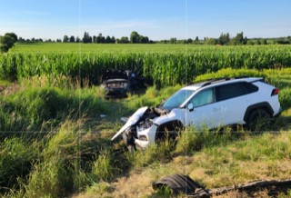 Damaged white SUV in tall grass, front end smashed, tire nearby