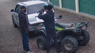 Man in a helmet and black shirt stands next to an ATV. A second man in a black shirt and jeans stands facing him, looking at a truck.
