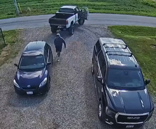 Two pickup trucks and a car parked on gravel. A person wearing a dark hoodie and sweatpants walks between the vehicles.