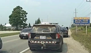 Police SUV with visible damage, parked on the shoulder of a road, other vehicles nearby, roadside sign reads "50 km/h Over Limit Max. Fine = $10 000 Roadside Suspension"