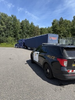 Black and white police SUV, partial view of a semi-truck and trailer in background, green trees.
