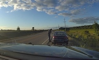 Person standing next to a black car on the shoulder of a road, grassy field, cloudy sky