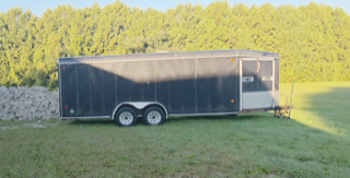 Black utility trailer with two axles and silver wheels, parked on green grass, trees in background