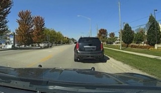 Black SUV ahead on a two-lane road, trees lining the street, clear blue sky