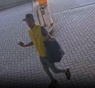 Man walking, carrying a black bag, wearing a yellow vest and a baseball cap.