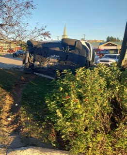 Damaged vehicle on its roof, foliage in foreground, street and buildings in background.