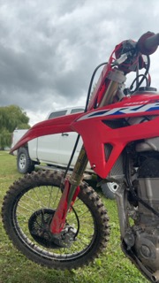 Red dirt bike with mud-caked tires and forks, parked on grass, pickup truck in background
