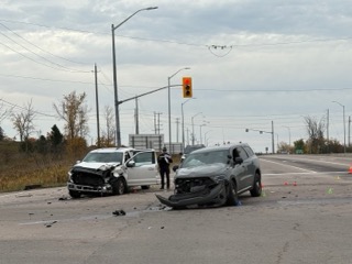 Two SUVs, heavily damaged, on a road. Debris scattered. Two people in dark clothing near the vehicles. Traffic signals visible. Trees and sky in background.