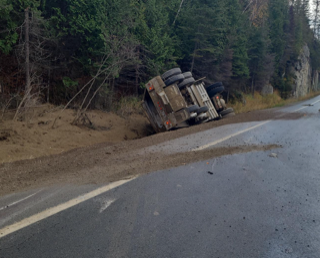 Semi-truck overturned on roadside, dense trees in background