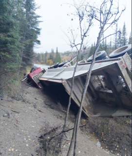 Tractor-trailer overturned on a gravel road, surrounded by trees.