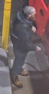 Man with a gray beard and dark jacket walking on a sidewalk.