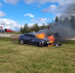 Car engulfed in flames on grass, black smoke rising