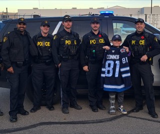 Five police officers and a child in a hockey jersey stand in front of a police vehicle.