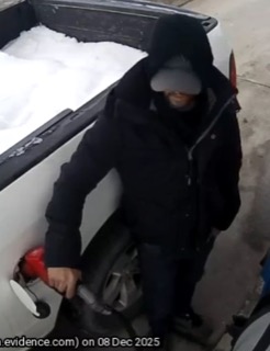 Person in a dark hooded jacket and baseball cap standing next to a pickup truck covered in snow.
