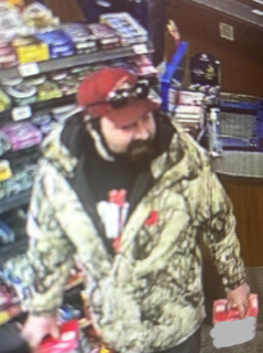 Man in camouflage jacket and red baseball cap, standing in a convenience store.