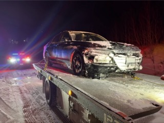 Damaged black sedan on a flatbed tow truck covered in snow.