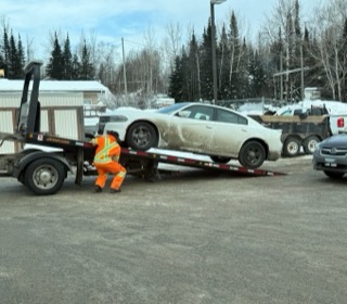 A silver sedan on a tow truck. A person in a yellow and orange safety vest is standing near the tow truck. Snow covers the ground.